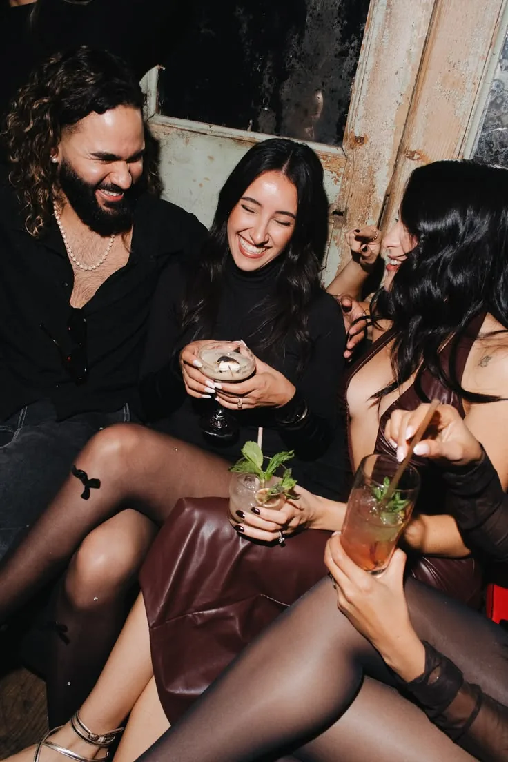 Three friends laughing on a bar banquette at night, cocktails in hand