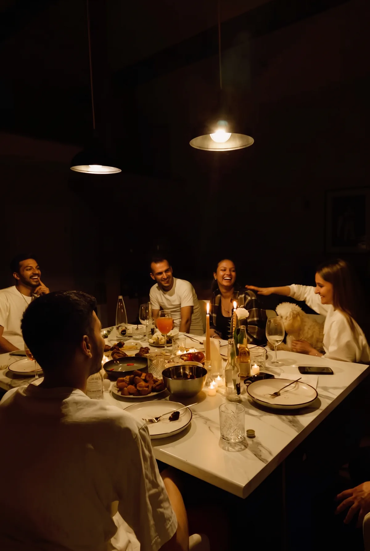 Six friends laughing around a candlelit marble table under two pendant lamps